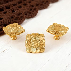 Może przedstawiać: Trzy złote gałki do szafek w kształcie kwiatów. Dwie gałki mają okrągły kształt, a jedna jest kwadratowa. Gałki znajdują się na białej drewnianej powierzchni z brązową szydełkową tkaniną w tle.