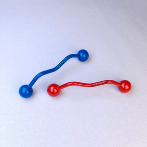 Op de afbeelding: Twee gebogen barbell-stijl lichaamsieraden, één in levendig blauw en één in helder rood. Elk stuk heeft gladde, afgeronde uiteinden en een golvende centrale staaf met het woord "VALDEX" duidelijk in het metaaloppervlak gegraveerd.
