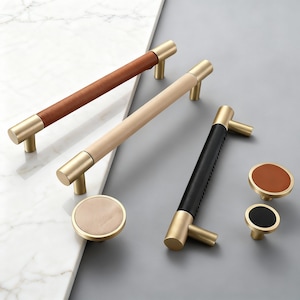 Op de afbeelding: Een set meubelbeslag met messing handgrepen en knoppen. De handgrepen hebben leren grepen in bruin, beige en zwart. De knoppen hebben bijpassende leren accenten in bruin en zwart. Afmetingen in centimeters.