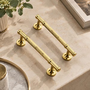 May include: Two gold-toned metal cabinet pulls featuring a textured, wood-grain-like pattern along the central bar. Each handle is supported by two circular mounting bases and includes a small, engraved logo on the side of the central bar.