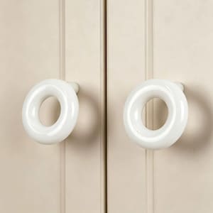 Op de afbeelding: Twee ronde, witte, glanzende kastknoppen met een gladde, ringvormige vorm. Elke knop heeft een opening in het midden en is gemonteerd op een kleine, cilindrische basis, wat zorgt voor een strak en reflecterend oppervlak op de deur.