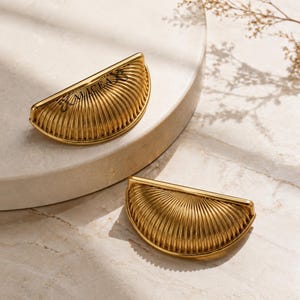 Puede incluir: Dos tiradores de cajón de metal dorado en forma de semicírculo con una textura acanalada en forma de abanico. Uno de los tiradores muestra el texto negro ALICEA grabado en su superficie. Estas piezas tienen una forma curva de concha.