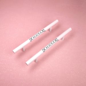 Puede incluir: Dos tiradores de armario cilíndricos de color blanco con dos puntos de montaje cada uno. La palabra ALICEA está impresa en letras mayúsculas negras en el centro de cada tirador, flanqueada por un símbolo similar al infinito a ambos lados.