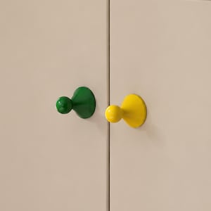 Op de afbeelding: Twee wandhaken bevestigd op een lichtbeige oppervlak. De ene haak is glanzend bosgroen met een afgerond knopuiteinde, en de andere haak is glanzend felgeel met een bijpassend afgerond knopuiteinde, beide voorzien van een conische basis.