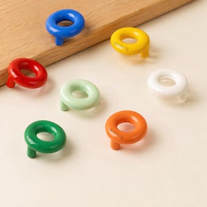 May include: A collection of seven circular cabinet knobs in various colours including blue, yellow, red, light green, white, dark green, and orange. Each knob features a smooth, glossy finish and a single mounting post on the underside.