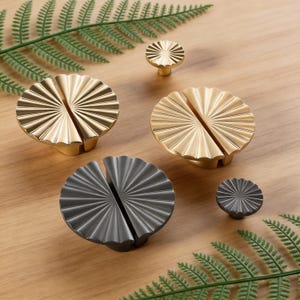 May include: A collection of decorative cabinet knobs in gold and dark gray. The knobs have a fan-like design with a split down the middle. Two sizes are shown, with the larger knobs split into two halves.