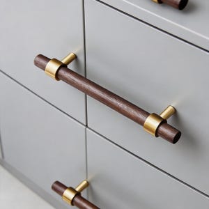 Pode incluir: Close-up de um armário cinza com puxadores de gaveta de madeira marrom escuro. Os puxadores têm um design cilíndrico com detalhes em metal dourado. O armário tem uma estética simples e moderna.