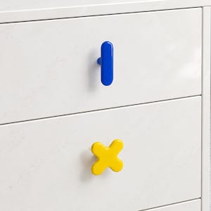 Puede incluir: Un mueble blanco de dos cajones con un acabado liso similar al mármol. El cajón superior tiene un pomo azul brillante en forma de píldora vertical, mientras que el cajón inferior presenta un pomo amarillo brillante en forma de cruz.