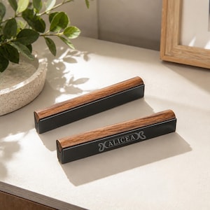 May include: Two rectangular display stands featuring a dark brown wood top piece with a visible grain pattern and a matte black base. The front-most stand features the engraved text ALICEA in a stylized font centered on the black metal surface.
