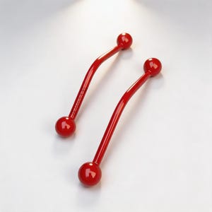 Könnte beinhalten: Zwei glänzend rote Schrankgriffe mit geschwungenem, länglichem Design. Jeder Griff hat eine glatte, einfarbig rote Oberfläche und endet an beiden Seiten in einem kugelförmigen Knopf, was für ein elegantes und einheitliches Aussehen sorgt.