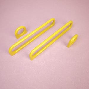 May include: Two bright yellow, elongated, U-shaped metal drawer pulls or cabinet handles, each featuring a long horizontal bar with a small, rounded loop at one end, presented as a pair of matching hardware components for furniture installation.