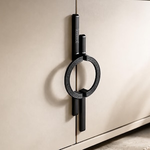 Può includere: Una maniglia per armadietto nero opaco con una base rettangolare verticale e un anello circolare con texture a coste. La maniglia è montata su un'anta di colore beige chiaro e presenta la scritta ALICEA incisa sulla barra verticale.