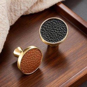 May include: Two decorative cabinet knobs with a gold finish. One knob features a black textured leather insert, and the other has a brown leather insert. The knobs are set on a dark wooden surface.