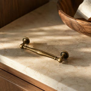 May include: A brass-toned metal drawer pull featuring a slender, contoured handle with rounded ends and circular mounting plates. The hardware has a weathered, antique-style finish with a smooth, slightly tapered center section and defined joints.