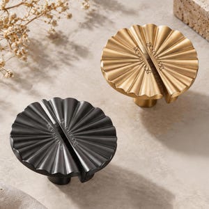 May include: Two circular cabinet knobs, one in a matte gold finish and one in a matte black finish. Each knob features a pleated, fan-like design with a central split and the brand name ALICEA engraved on the surface of the metal hardware.