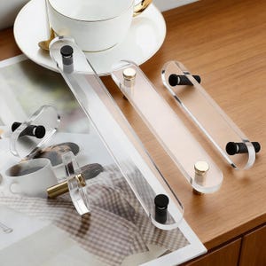 Könnte beinhalten: Klare Acryl-Schrankgriffe mit schwarzen und goldenen Akzenten. Die Griffe sind rechteckig mit abgerundeten Enden und werden mit Schrauben befestigt. Die Hardware wird auf einer Holzoberfläche mit einer Tasse und Untertasse im Hintergrund präsentiert.