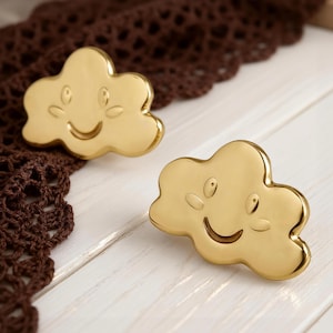Puede incluir: Dos tiradores dorados en forma de nube con caras sonrientes. Los tiradores están sobre una superficie de madera blanca con un ribete de encaje de ganchillo marrón. Cada tirador tiene una cara sonriente con ojos y mejillas.