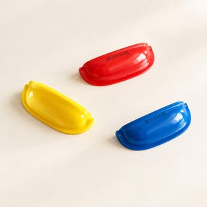 以下が含まれることがあります： 鮮やかな赤、黄、青の半円形カップ型引き出し用取っ手3点。光沢のある各ハンドルは滑らかな曲線を描いており、前面にはブランド名「ALICEAX」が黒い文字で刻印されています。