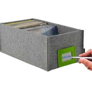 Puede incluir: Una caja de almacenamiento de fieltro gris con una etiqueta verde que dice "DVDs". La caja está llena de DVD.