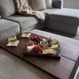 May include: A rectangular dark wood serving tray featuring a charcuterie arrangement with red grapes, sliced salami, brie, hard cheese wedges, fresh figs, almonds, crackers, and a small bowl of honey with a wooden dipper, plus two glasses of white wine.