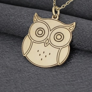 Könnte beinhalten: Eine goldfarbene Eulen-Anhänger-Halskette. Der Anhänger zeigt ein detailliertes Eulen-Design mit großen Augen und einem strukturierten Körper. Die Halskette ist zart und goldfarben, vor grauem Hintergrund.