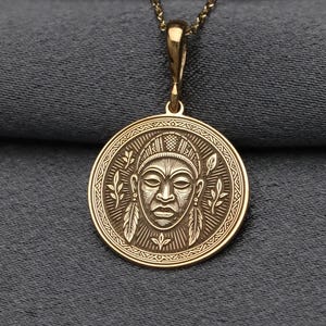 May include: Gold-colored pendant necklace featuring a circular pendant with a detailed face design. The pendant is suspended from a delicate chain. The face design is surrounded by a border of leaves and feathers. The necklace is displayed against a gray background.