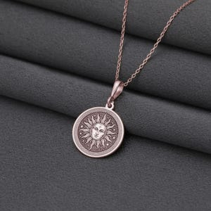 A round pendant with a sun design is displayed on a chain against a gray fabric background.