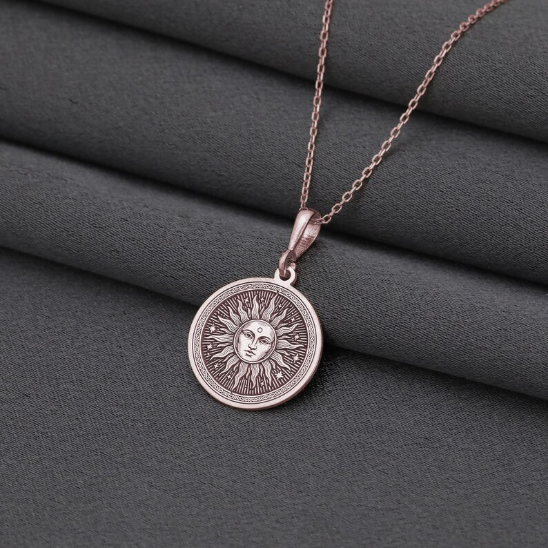 A round pendant with a sun design is displayed on a chain against a gray fabric background.
