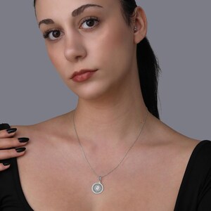 A woman with dark hair wearing a black top and a silver necklace with a pendant is shown in a close-up portrait against a gray background.