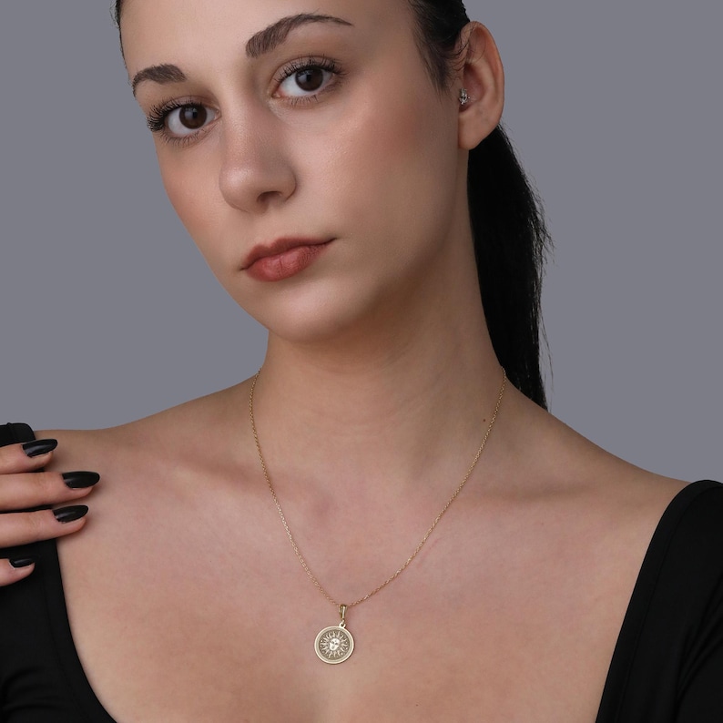 A woman with dark hair wearing a black top and a gold necklace with a pendant is shown in a close-up portrait against a gray background.