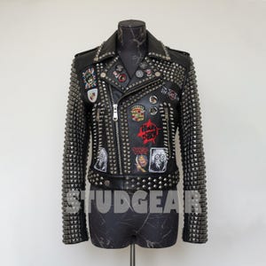 May include: Black leather jacket with silver studs and patches. The jacket features a classic biker style with a notched collar, zipper closure, and multiple decorative patches. The sleeves and shoulders are heavily studded. The text "STUDGEAR" is visible.