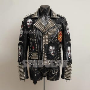 May include: Black leather jacket with silver metal studs and patches featuring faces. The jacket has a notched collar, a zipper closure, and a belt at the waist. The sleeves and collar are heavily studded.