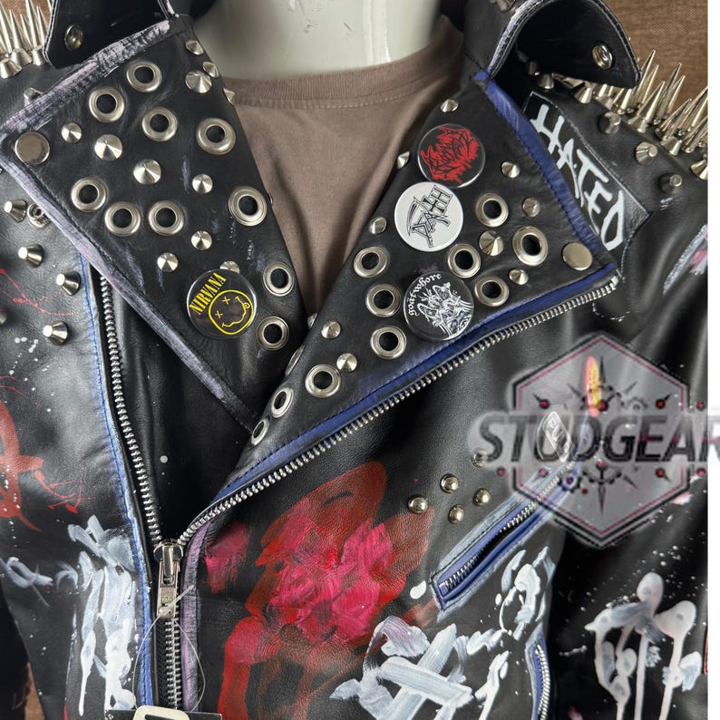 May include: A black leather jacket featuring silver metal studs, spikes on the shoulders, and hand-painted red and white abstract designs. The lapels are decorated with circular pin-back buttons, including one with the Nirvana logo and text.