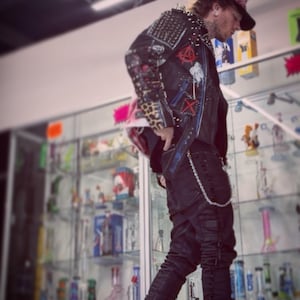 May include: A black leather jacket adorned with silver metal studs, red and white patches, and leopard print accents on the sleeves, paired with black trousers featuring a silver chain accessory and a black baseball cap with a red patterned brim.