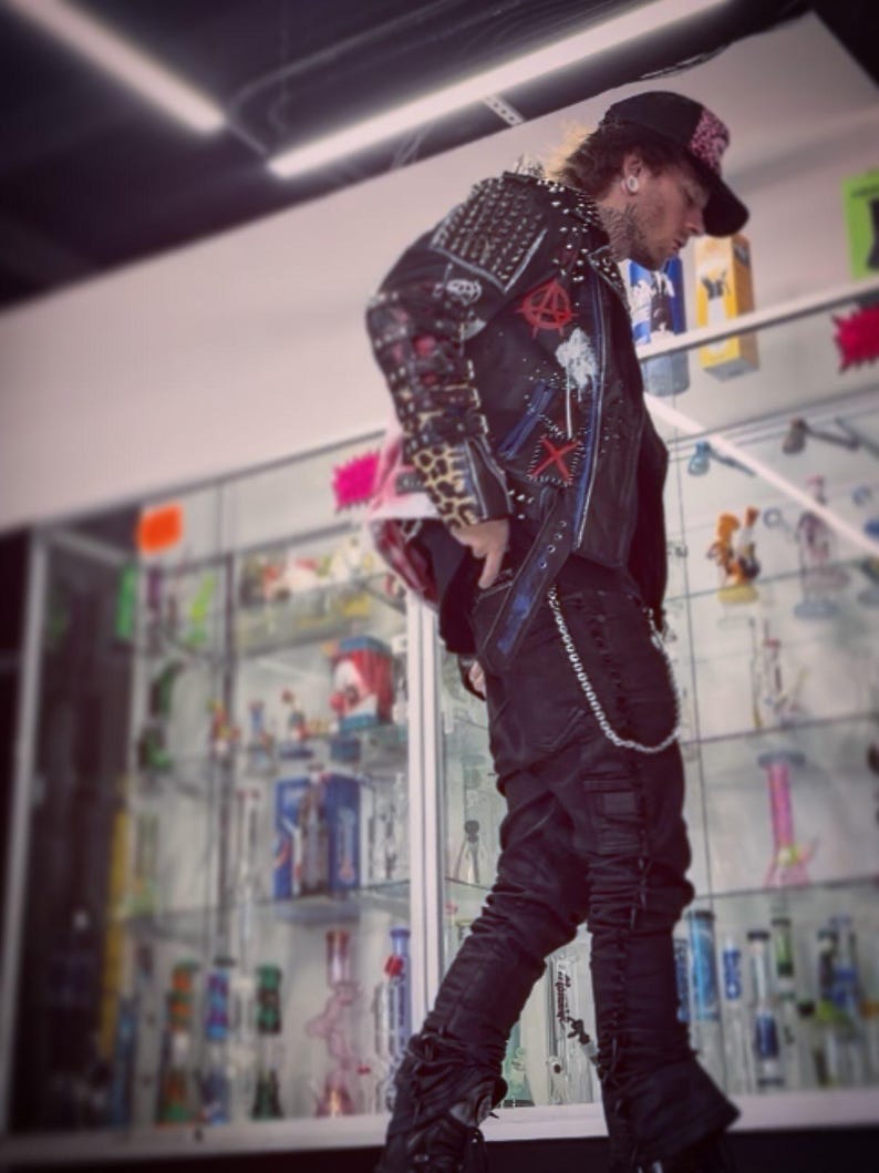 May include: A black leather jacket adorned with silver metal studs, red and white patches, and leopard print accents on the sleeves, paired with black trousers featuring a silver chain accessory and a black baseball cap with a red patterned brim.