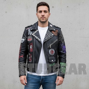 May include: Black leather jacket with silver studs on the shoulders and sleeves. The jacket features various patches with graphic designs and text. The model is wearing a white t-shirt and blue jeans. The jacket has a zipper closure and multiple pockets.