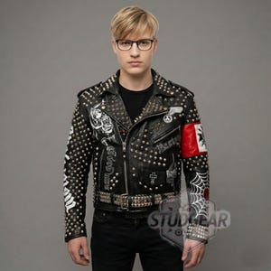 Könnte beinhalten: Schwarze Lederjacke mit silbernen Nieten, Aufnähern und grafischen Designs. Mit einem Nietengürtel und einer rot-weißen Armbinde. Die Jacke hat einen Reißverschluss und ist ein klassischer Biker-Stil.