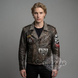 Op de afbeelding: Bruine leren jas met zilveren studs op de kraag en schouders. De jas heeft patches met schedel- en tekstontwerpen. Het heeft een ritssluiting en een riem. De tekst "STUDGEAR" is rechtsonder zichtbaar.