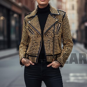 Puede incluir: Chaqueta de cuero negra adornada con tachuelas doradas. La chaqueta presenta un estilo motero clásico con cuello de solapa, cremallera asimétrica y múltiples bolsillos. La modelo lleva un cuello alto negro y vaqueros negros.