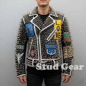 Può includere: Giacca di pelle nera con borchie argentate e toppe decorative. Presenta un bordo bianco, una chiusura a zip e la parola "LOVE" su una toppa gialla. Altre toppe includono un ragno, un quadrato blu con borchie e la parola "PIXIES".