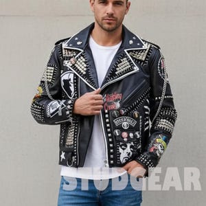 May include: Black leather jacket with silver studs, patches, and pins. The jacket has a white collar and zipper detail. Patches include a graphic of a person, a globe, and the text "Ashley Cure". The jacket is from Studgear.