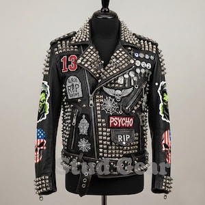 Op de afbeelding: Zwarte leren jas rijkelijk versierd met zilveren studs en spikes. Voorzien van patches met het nummer 13, een grafsteen, een spinnenweb en het woord "PSYCHO". Inclusief een riem met een zilveren gesp en studs. Een gedurfde, edgy mode-uiting.
