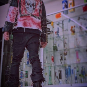 May include: A black jacket featuring a large skull graphic on the back, red distressed fabric accents, and silver metal studs along the shoulders and sleeves. The jacket is paired with black trousers and black boots with a metallic drip design.
