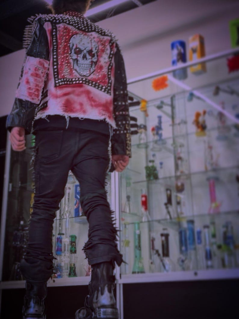 May include: A black jacket featuring a large skull graphic on the back, red distressed fabric accents, and silver metal studs along the shoulders and sleeves. The jacket is paired with black trousers and black boots with a metallic drip design.