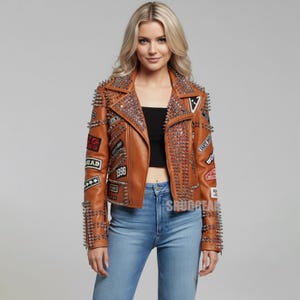 May include: Brown leather jacket with silver spikes and patches. The jacket has a classic biker style with a notched collar, a front zipper, and multiple decorative patches. The jacket is a medium brown color.