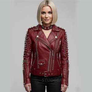 May include: A burgundy leather jacket with silver metal studs and zips. The jacket has a classic biker style with a notched collar, asymmetrical zip closure, and a belt at the waist. The sleeves and shoulders are heavily studded.