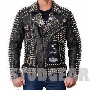 Puede incluir: Chaqueta de cuero negro con tachuelas de metal plateado. La chaqueta tiene múltiples cremalleras, parches con logotipos de bandas y un cuello tachonado. Un estilo motero clásico.