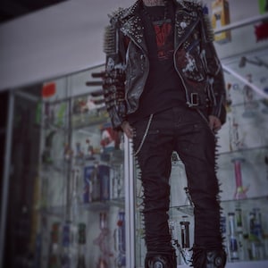 May include: A black leather jacket adorned with numerous metal spikes and studs, worn over a black t-shirt with a red graphic print. The outfit includes black trousers with side spikes and a silver chain, paired with black platform boots and a cap.