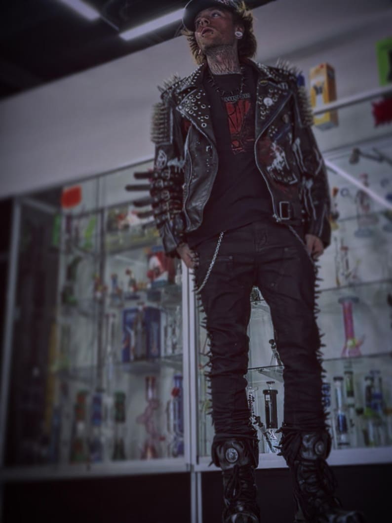 May include: A black leather jacket adorned with numerous metal spikes and studs, worn over a black t-shirt with a red graphic print. The outfit includes black trousers with side spikes and a silver chain, paired with black platform boots and a cap.