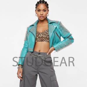 Op de afbeelding: Een turquoise leren jas met zilveren spikes op de schouders en kraag. De jas heeft een ritssluiting en wordt gedragen met een top met luipaardprint en een grijze broek. Het woord "STUDGEAR" staat op de afbeelding.